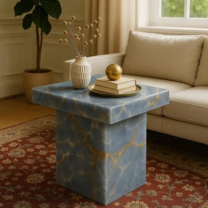 May include: A square, blue side table with gold veining. The table has a solid, block-like base and a flat top. A gold sphere and books rest on top. A vase with decorative stems sits nearby.