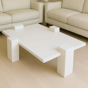 Può includere: Un moderno tavolino rettangolare bianco con quattro gambe a blocchi quadrati e quattro blocchi più piccoli sopra. Il tavolo è in una stanza con due divani color crema e un pavimento chiaro.