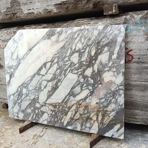 Puede incluir: Una gran losa rectangular de m&aacute;rmol con un patr&oacute;n veteado blanco y gris. La losa est&aacute; sostenida por un soporte met&aacute;lico y se apoya contra una pila de otros bloques de piedra. El m&aacute;rmol tiene una superficie pulida.