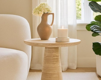 May include: A beige, round side table with a textured, conical base supports a vase of white flowers and a lit candle. The vase is a light brown color with a handle. The candle sits on a small, round, white base. A white, textured rug is on the floor.