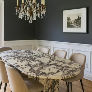 May include: An oval marble dining table with a dark veined pattern, surrounded by six beige upholstered chairs with black legs. A vintage chandelier hangs above the table. The room has dark gray walls, white trim, and a framed landscape print.