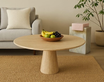 May include: A round, light wood coffee table with a pedestal base. A wooden bowl of fruit sits on top, including bananas, green apples, and grapes. A beige sofa and a woven rug are in the background.