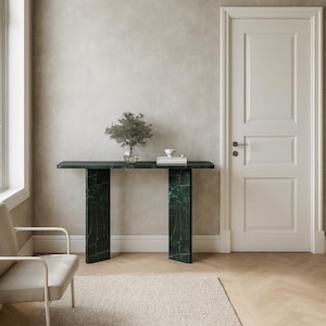 May include: A dark green marble console table with a rectangular top and two vertical supports. The table is decorated with a small plant in a glass vase and a stack of white books. The room has a neutral color palette.