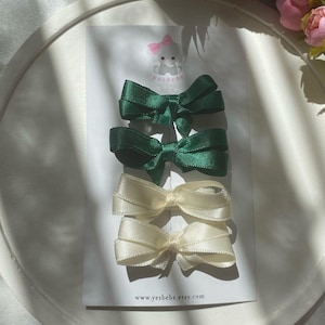 May include: Four satin hair bows in two colors: dark green and cream. The bows are arranged on a white card with the text "yesbebe.etsy.com" at the bottom. The top of the card has a logo of a pink bow.