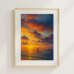 Op de afbeelding: Ingelijste kunstdruk met een persoon die bij zonsondergang aan het suppen is. De lucht is een mix van oranje, geel en blauw. Het kunstwerk is ingelijst in een lichte houttint.