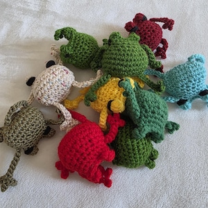 Può includere: Una collezione di rane all'uncinetto fatte a mano in vari colori, tra cui verde, rosso, giallo e blu. Le rane hanno dettagli e sono disposte su una superficie bianca. Queste creature stravaganti sono perfette per un giocattolo per bambini o un oggetto di arredamento unico.