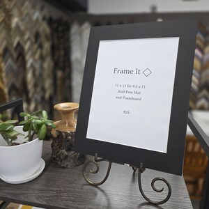 May include: A black picture frame with a white mat, displayed on a decorative stand. The frame holds a white paper with the text "Frame It" and details about the mat and foamboard. The frame is 11 x 14 inches for an 8.5 x 11 inch image.