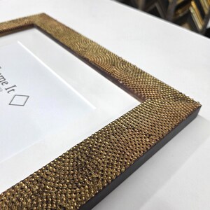 May include: Close-up of a rectangular picture frame with a textured, gold-colored border. The frame has a white mat and a dark brown inner edge. The text "Frame It" is visible on the white mat.