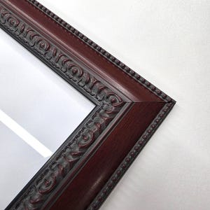 May include: Close-up of a rectangular picture frame with a dark burgundy finish. The frame has ornate, raised detailing along the inner and outer edges, with a beaded border. The frame's corner is visible against a white background.
