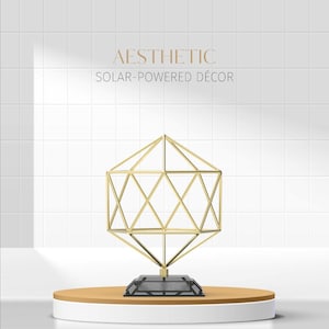 May include: Aesthetic solar-powered decor. A geometric gold-colored sculpture with a diamond-like shape sits on a round, two-tiered stand. The stand has a white base and a light brown top. The sculpture is mounted on a black base. The background is white with a tiled pattern.