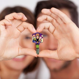 Puede incluir: Una llave decorativa con una imagen de dibujos animados de una reina con una corona morada y dorada. La llave tiene una hoja plateada y una franja verde cerca de la parte inferior. La llave se sostiene entre dos manos.