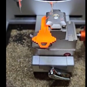 Puede incluir: Una m&aacute;quina de cortar llaves con una llave naranja sujeta. La m&aacute;quina es plateada y gris, con una broca colocada encima de la llave. La llave es de color naranja brillante y est&aacute; siendo cortada por la m&aacute;quina.