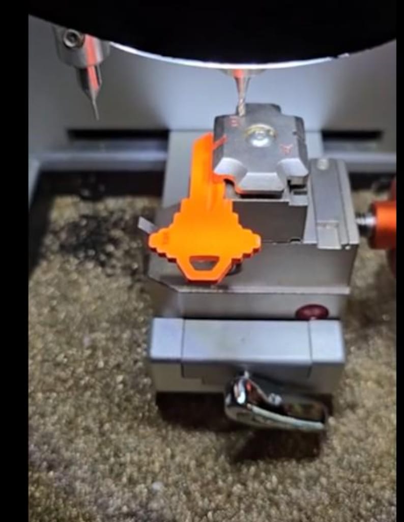 Puede incluir: Una m&aacute;quina de cortar llaves con una llave naranja sujeta. La m&aacute;quina es plateada y gris, con una broca colocada encima de la llave. La llave es de color naranja brillante y est&aacute; siendo cortada por la m&aacute;quina.