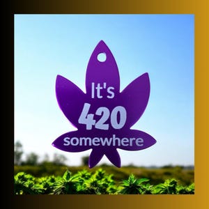 May include: A purple cannabis leaf-shaped tag with the text "It's 420 somewhere" in white. The tag has a hole at the top and is set against a blue sky and green plants.