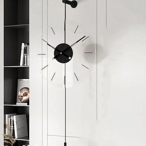 Könnte beinhalten: Eine moderne Wanduhr mit transparentem Zifferblatt und schwarzen Zeigern und Akzenten. Die Uhr hat ein kreisförmiges Design mit minimalistischen Stundenmarkierungen und einem Pendel. Die Uhr ist an einer weißen Wand befestigt.