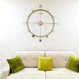 Puede incluir: Un gran reloj de pared de metal dorado con números romanos y un diseño minimalista cuelga sobre un sofá color crema. Tres cojines cuadrados verdes y amarillos añaden un toque de color a la sala de estar de tonos neutros.