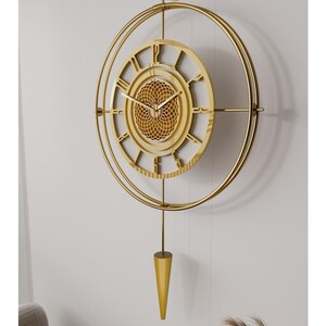 Könnte beinhalten: Eine goldfarbene, dekorative Wanduhr mit Pendel. Die Uhr hat römische Ziffern und ein detailliertes, kreisförmiges Design. Die Uhr ist an einer weißen Wand befestigt und hat ein goldfarbenes Pendel, das nach unten hängt. Der Durchmesser der Uhr beträgt ca. 46 cm.