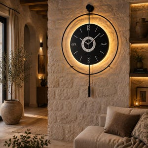 May include: A circular black wall clock featuring a central dial with white numerals at 12, 3, 6, and 9 o'clock. The clock is framed by a thin black metal ring and includes a vertical pendulum arm with a cylindrical weight and integrated backlighting.