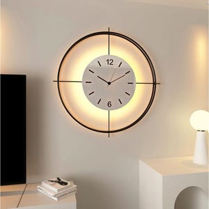Puede incluir: Un reloj de pared moderno con un marco de metal negro y un diseño circular. La esfera del reloj es blanca con manecillas y marcadores de hora negros. El reloj tiene una luz cálida y brillante detrás de la esfera.
