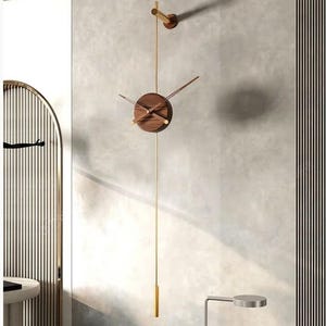 May include: A modern wall clock with a round, dark wood face and gold-colored hands. The clock hangs from a gold-colored rod, with a matching weight at the bottom. A smaller, similar design is mounted above the clock. The clock is approximately 12 inches in diameter.