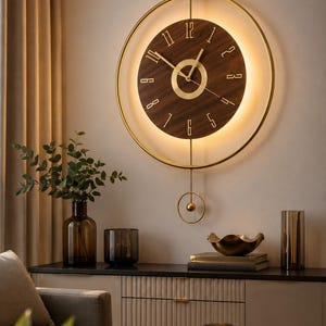 Op de afbeelding: Een ronde wandklok met een wijzerplaat van donkerbruin hout, gouden cijfers en wijzers. De klok is omvat door een dunne goudkleurige metalen ring met een geïntegreerde lichtbron en een gouden slinger die onderaan in het midden hangt.