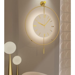 Könnte beinhalten: Eine moderne Wanduhr mit einem goldfarbenen kreisförmigen Rahmen und einem weißen Zifferblatt. Die Uhr hat goldene Zeiger und Stundenmarkierungen. Das Design umfasst ein Pendel und einen kleinen, runden Akzent oben. Die Uhr ist an einer neutralfarbenen Wand befestigt.