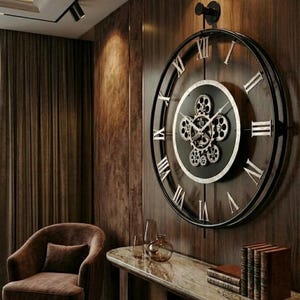 May include: A large, circular wall clock with a black metal frame and Roman numerals. The clock face is black with silver gears at the centre. The clock is mounted on a wood-panelled wall, with a brown armchair and a marble-topped table in the foreground.
