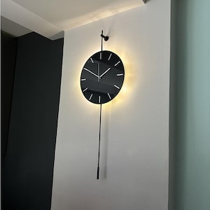 May include: A modern black wall clock with a circular face and a pendulum. The clock has a minimalist design with white hour markers and hands. The clock is illuminated with a warm light, creating a halo effect.