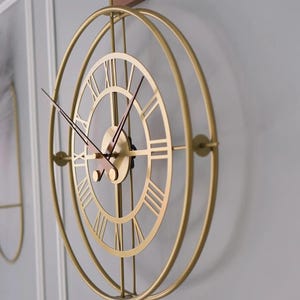Op de afbeelding: Een goudkleurige metalen wandklok met Romeinse cijfers. De klok heeft een cirkelvormig ontwerp met twee concentrische ringen en een houten montagebeugel. De wijzers zijn zwart en de secondewijzer is rood.