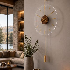 May include: A wall clock featuring a circular transparent acrylic face with gold-toned metal hour markers and hands. The center is a dark brown wood disc. A vertical gold metal rod extends above and below the face, ending in a cylindrical weight.