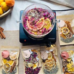 Puede incluir: Una olla de vidrio sobre una placa de cocina negra llena de rodajas de cítricos secos, pétalos de rosa y hierbas. Varias bolsas selladas de cítricos, hierbas y especias secas están dispuestas alrededor de la olla. Un cuenco de madera con naranjas y limones está en el fondo.