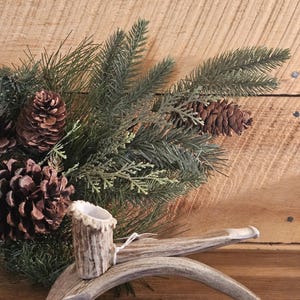 May include: A rustic decorative piece featuring a small, cylindrical holder with a textured surface, possibly for a candle, set on a curved antler base. The arrangement includes pinecones and evergreen branches, creating a natural, woodland theme.
