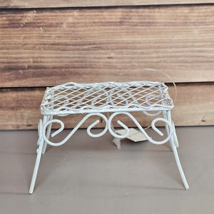 Può includere: Un piccolo supporto per piante in metallo bianco con un design decorativo a volute. Il supporto ha una parte superiore quadrata con un motivo a griglia e quattro gambe angolate. Il supporto è alto circa 15 cm.