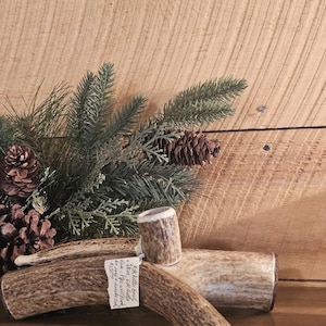 May include: A rustic arrangement featuring elk antler pieces, including a small cup and sections of antler. The antlers are a natural brown color, with a rough texture. Green pine branches and pine cones add a natural touch. The background is a wooden surface.