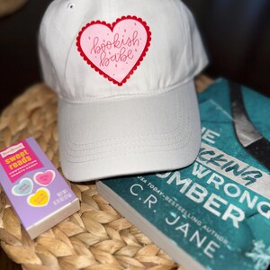 May include: A white baseball cap with a pink heart-shaped graphic that reads "bookish babe." Beside the cap are a book and a box of conversation heart candies. The book has a teal cover and the title "Picking the Wrong Number."