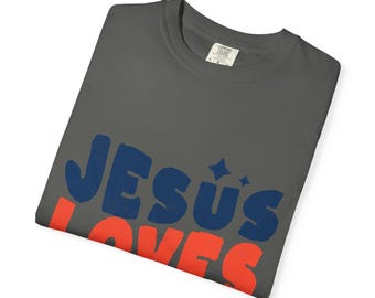 May include: A dark gray t-shirt with the words "JESUS LOVES" in blue and red block letters. The shirt has a crew neck and a tag on the inside collar. The text is in a retro, playful font.
