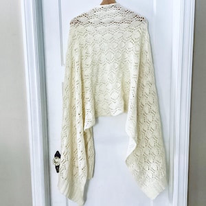 Può includere: Sciarpa color crema a trama aperta con un abbellimento floreale. La sciarpa è drappeggiata su un appendiabiti in legno, mettendo in risalto l'intricato motivo a maglia e il design fluido. La texture delicata e il colore neutro lo rendono un accessorio versatile.