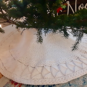 May include: A white knitted Christmas tree skirt with a scalloped edge and a delicate cable knit pattern.