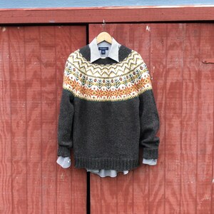 Può includere: Un maglione marrone con un motivo geometrico colorato sulla spalla. Il maglione ha un collo rotondo e maniche lunghe.
