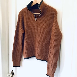 Puede incluir: Un suéter marrón texturizado con cierre de media cremallera y cuello negro. El suéter está colgado en una percha de madera contra una puerta blanca. Los puños y el dobladillo tienen un ribete negro.