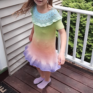 May include: A young girl wearing a rainbow ombre knit dress with a crocheted lace collar. The dress has a ruffled hem and is a light green, orange, and purple color.