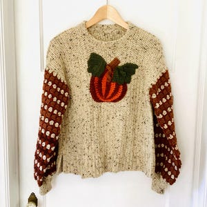 May include: A beige knit sweater with a large pumpkin applique. The pumpkin is orange and red with green leaves. The sleeves are brown with white square patterns. The sweater is hanging on a wooden hanger.