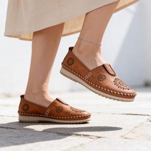 Woman wearing handmade tan leather slip-on flats with floral cutout detailing, walking outdoors in soft sunlight with a cream skirt and silver anklet.