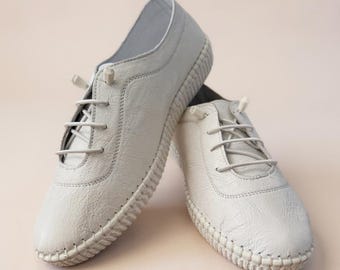 May include: A pair of off-white, textured leather shoes with matching laces. The shoes have a rounded toe and a stitched rubber sole. The laces are tied, and the shoes are displayed on a light pink background.