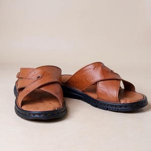 Puede incluir: Un par de sandalias de cuero marr&oacute;n con un dise&ntilde;o de correas cruzadas. Las sandalias tienen una suela negra y un cierre de hebilla en una sandalia. El cuero parece desgastado y texturizado, lo que sugiere un estilo vintage.