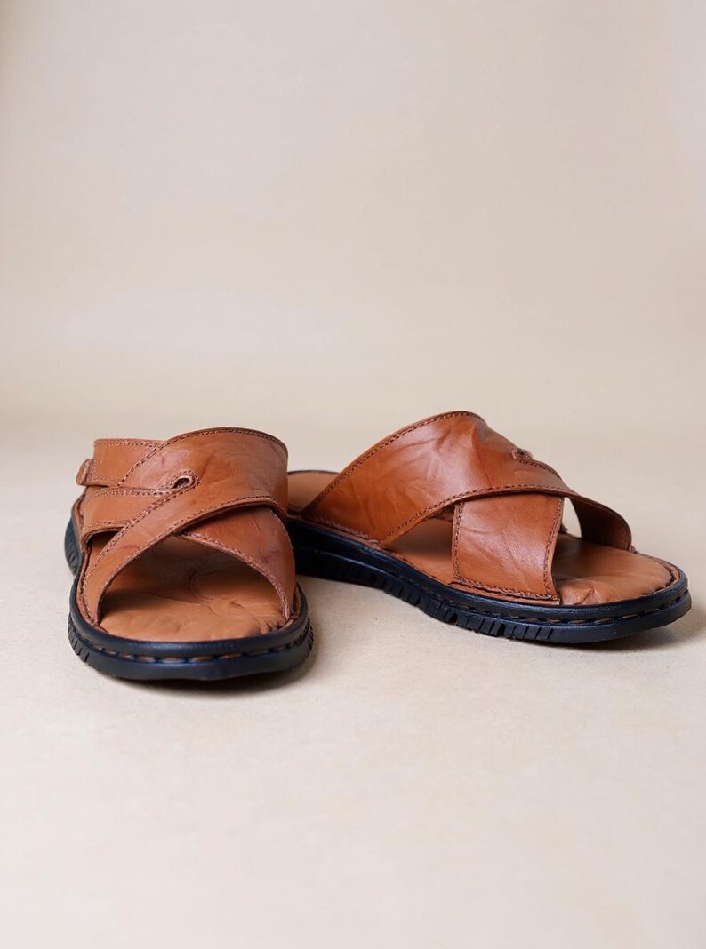 Puede incluir: Un par de sandalias de cuero marr&oacute;n con un dise&ntilde;o de correas cruzadas. Las sandalias tienen una suela negra y un cierre de hebilla en una sandalia. El cuero parece desgastado y texturizado, lo que sugiere un estilo vintage.