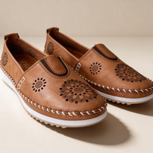 Pair of handmade tan leather slip-on flats with floral cutout design and white soles, photographed at an angle on a clean beige studio background.