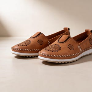 Handmade tan leather slip-on flats placed side by side on a soft beige background, highlighting the side profile, stitching, and floral cutout details.
