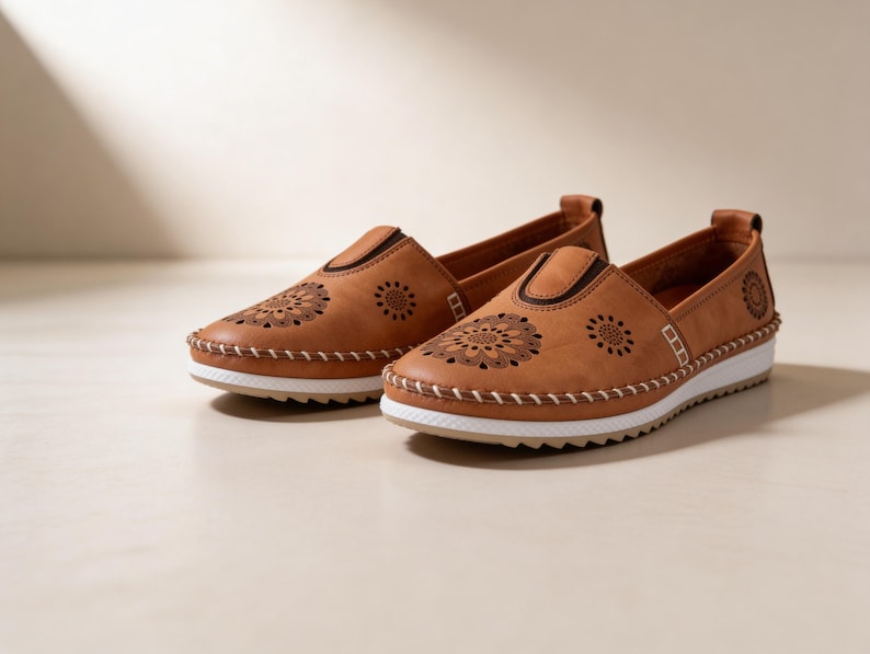 Handmade tan leather slip-on flats placed side by side on a soft beige background, highlighting the side profile, stitching, and floral cutout details.