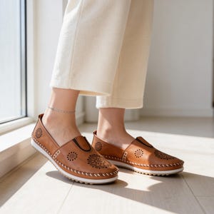 Woman sitting by a window wearing handmade tan leather slip-on flats with floral cutout details, paired with light-colored pants in natural indoor light.